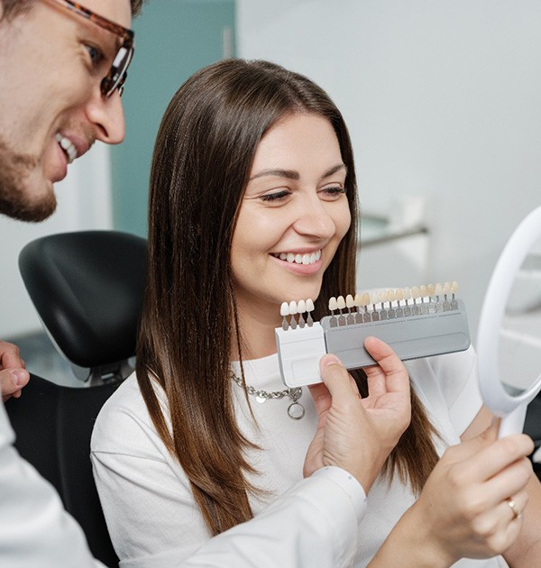 Dentist holding shade guide to woman’s teeth as she looks in mirror